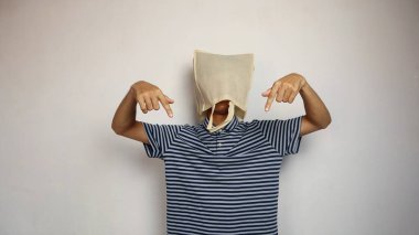 Kafasında alışveriş torbası olan genç bir Asyalı adam el hareketi ile izole edilmiş beyaz bir arkaplanı işaret ediyor.