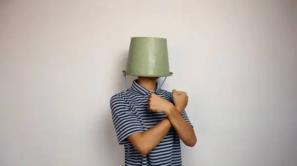 young man using a bucket on his head on a white background. showing a crossed arms gesture as if saying no. funny and strange moments