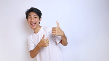 Heyecanlı, beyaz tişörtlü, komik suratlı Asyalı genç adam başparmağını kaldırıyor. Tamam, harika, sakin, izole edilmiş beyaz arka plan kazanıyor.