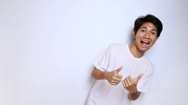 Heyecanlı, beyaz tişörtlü, komik suratlı Asyalı genç adam başparmağını kaldırıyor. Tamam, harika, sakin, izole edilmiş beyaz arka plan kazanıyor.