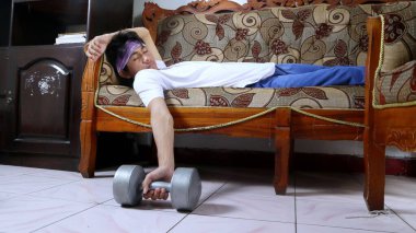 Genç adam dambılları tutarken kanepede uyuyakaldı. Egzersiz yapmaktan yoruldum. izole edilmiş beyaz arkaplan