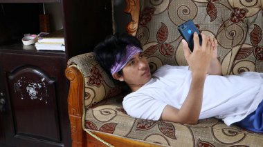 Genç Asyalı adam akıllı telefonunu kullanarak kanepede uzanıyor.