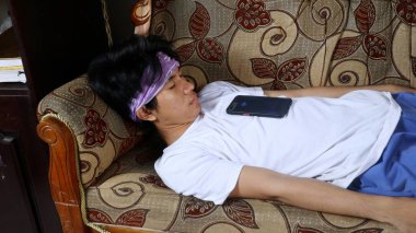 young asian man sleeping on sofa with smartphone on his chest clipart
