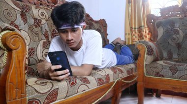 Genç Asyalı adam akıllı telefonunu kullanırken kanepede uzanıyor.