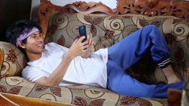 Mutlu Asyalı genç adam akıllı telefonunu kullanırken kanepede uzanıyor.