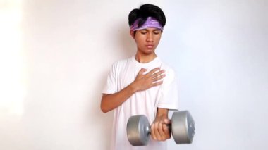 Beyaz tişörtlü genç bir Asyalı egzersiz yapıyor. Kol kaslarını çalıştırmak için halterlerini kaldırıyor. Göğsünü tutuyor.