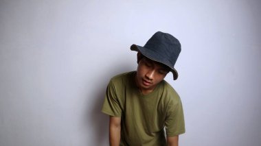 Şapkalı genç Asyalı adam uyuşuk, yorgun, kamburlaşmış, soyutlanmış beyaz bir arka planı var.