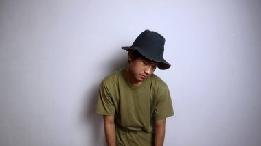 Şapkalı genç Asyalı adam uyuşuk, yorgun, kamburlaşmış, soyutlanmış beyaz bir arka planı var.