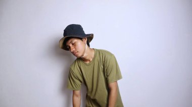 Şapkalı genç Asyalı adam uyuşuk, yorgun, kamburlaşmış, soyutlanmış beyaz bir arka planı var.