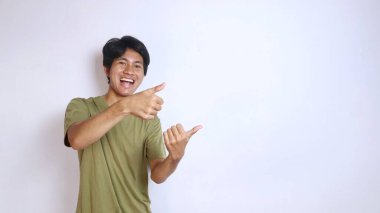 Heyecanlı genç Asyalı adam, izole edilmiş beyaz bir arka planda baş parmağını kullanarak kenarı işaret ediyor.