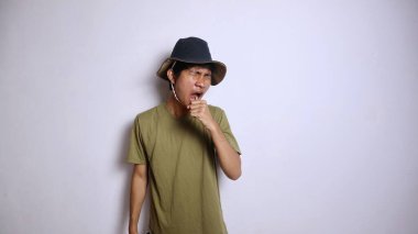 Şapkalı genç Asyalı adam yorgun, uykulu, esneyen hareketlerle ağzını izole edilmiş beyaz arka planla kapatıyor.