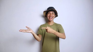 Heyecanlı Asyalı genç adam, izole edilmiş beyaz arka planda boş avuç içlerine işaret eden bir şapka takıyor.