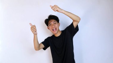 Siyah tişörtlü heyecanlı genç Asyalı adam, iki elinde baş parmaklarıyla izole edilmiş beyaz bir arkaplanda işaret veriyor. kopya alanı, başarı, mutluluk, evet de, tamam, güzel, lezzetli