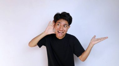 Asyalı heyecanlı genç bir adam, siyah bir gömlek giyiyor ve elleri kulaklarının yanında, avuçları açık, izole beyaz bir arka planda hareket ediyor. Boşluğu kopyala, bir şey dinle