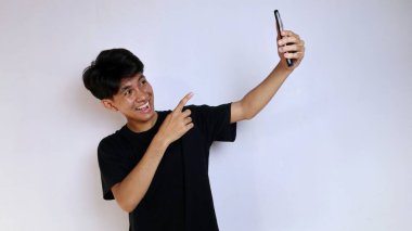 Siyah tişört giyen mutlu Asyalı genç adam izole edilmiş beyaz arka planda akıllı telefon kamerasıyla selfie çekiyor.