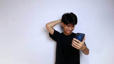 Siyah tişört giyen kafası karışmış genç Asyalı adam izole edilmiş beyaz arka planda akıllı telefon tutuyor.