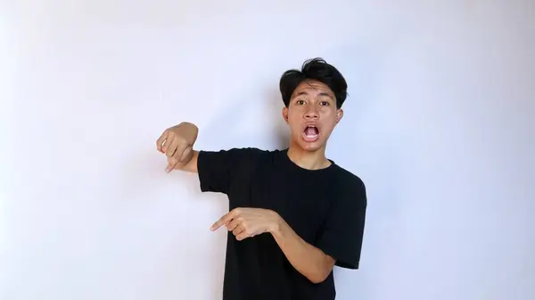 stock image shocked young asian man wearing black shirt gesturing with hand pointing down on isolated white background. copy space