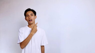 Şok olmuş Asyalı genç erkek beyaz gömlekli el hareketleriyle sağ tarafı işaret ediyor ve izole edilmiş beyaz arka planla ileriye bakıyor.