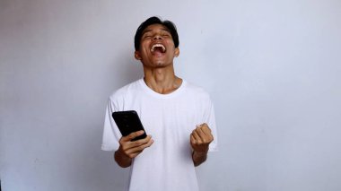 Asyalı genç adam beyaz gömlek giyip, beyaz arka planlı akıllı telefon tutarak bağırmaktan mutlu oluyor.