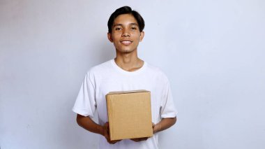Asyalı genç adam beyaz gömlek içinde gülümsüyor. Elinde beyaz arka planda bir paket kutusu var.