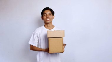 Asyalı genç adam beyaz gömlek içinde gülümsüyor. Elinde beyaz arka planda bir paket kutusu var.