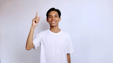 Beyaz tişörtlü genç Asyalı adam izole edilmiş beyaz arka plana işaret ederken gülümsüyor, boşluğu kopyala