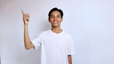Beyaz tişörtlü genç Asyalı adam izole edilmiş beyaz arka plana işaret ederken gülümsüyor, boşluğu kopyala