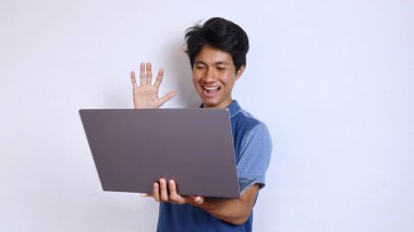Asyalı genç adam gülümsüyor, el kol hareketleri yapıyor ve dizüstü bilgisayarına bakıp beyaz arka planda merhaba diyor.
