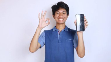 happy asian young man gestures okay while holding smartphone with isolated white background clipart