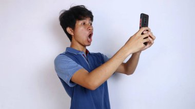 Şok olmuş Asyalı genç adam hareketi kaldırıyor ve izole beyaz arka plana sahip akıllı telefona bakıyor.