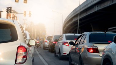 Rear side view of white car with turn on brake light. Put a traffic signal forbidden to pass in the intersection with cars parked in a queue. Blurred view of a concrete bridge under the everning sky.