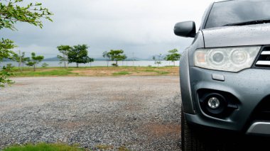 Yağmur damlalarıyla parktaki sisli gri araba farlarına odaklan. Bahçedeki toprak ve çakıl yolunda park etmek. Yağmurdan sonra bulutlu gökyüzünün altında nehir ve dağın arkaplanı.