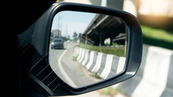 Mirror wing of car cover of black color. inside glass mirror with  curves road and barrier platforms with other cars driving on the road at day. and beside with bridge.
