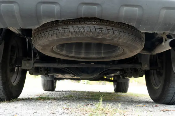 Maintenance background of car's suspension reveals the rear drive shaft. Cars parked on the soil road with gravel.
