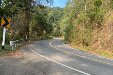 Dağların yanındaki eğri asfalt yolda. Kıvrımlar için trafik işaretleri. Yol kenarında çimenler ve yeşil ağaçlarla çevrili. Mavi gökyüzünün altındaki dağ tabanı.