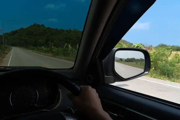 View from the inside of a car. View from the driver\'s seat. Driving on the asphalt road with nature of tree and mountain beside road. Under blue sky in the day time.