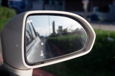 Arabanın beyaz renkli aynanın yan tarafında. Arka planda dikiz aynası var. Gökyüzünün altında yeşil bir bahçe var..