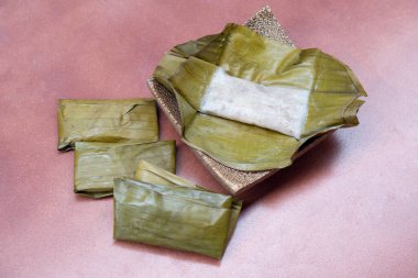 Muz yaprağında buğulanmış muzlu kek. Hindistan cevizi palmiyesi desenli bir tabağa yerleştirilmiş. Paketlenmiş muzlu kekler yan yana dizilmiş. Kırmızı döşemeye yerleştirilmiş..