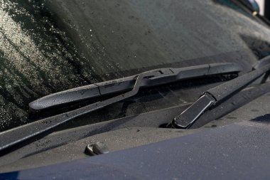 Arabanın ön cam sileceğine yakın çekim. Silecek bıçağı siyah ve kauçuktan yapılmış gibi görünüyor. Silecek kolu da siyahtır ve üzerinde metal bir yay vardır. Arka plan bulanık, arabanın kaputu..