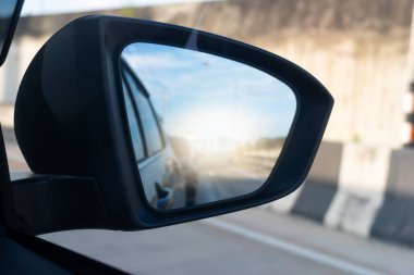 Bir arabanın dikiz aynası. Arabanın sürücü tarafındaki kapısına monte edilmiş. Aynanın yüzeyi biraz renkli ve yolu yansıtıyor. Gökyüzü yansımanın üst köşesinde görünür..