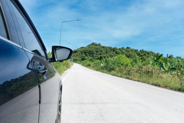 Asfalt yolda, koyu renkli arabanın yanında. Parlak ışık yola yansıyor. Yolun her iki tarafı da yeşil ağaçlar, çimenler ve aydınlatma direkleriyle dolu. Mavi gökyüzünün altındaki tepelere doğru gidiyor..