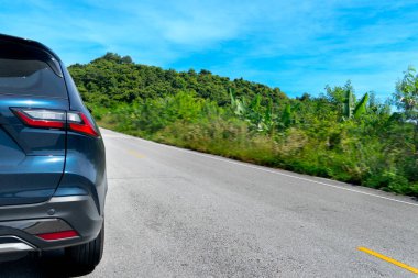 Asfalt yolda giden koyu mavi arabanın arka tarafında. Yolun her iki tarafı da yeşil ağaçlar, çimenler ve aydınlatma direkleriyle dolu. Mavi gökyüzünün altındaki tepelere doğru gidiyor..