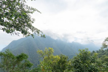Alçak asılı bulutlarla kısmen gizlenmiş ve ön plandaki yeşil yapraklarla çerçevelenmiş sisli dağ sıraları. Pha Bong 'un tarihi tepeleri. Tayland 'dan Mae Hong Oğlu.