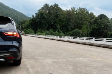 Dark blue of SUV is parked on a bridge with a mountain and trees in the background. Car turn light signal. The bridge stretches into the distance. with blue sky. Traveling in Thailand Mae Hong Son. clipart