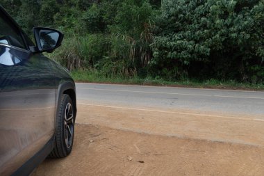 Koyu renkli bir arabanın bir parçası kaldırımlı bir yolun yanındaki toprak bir omzun üzerine park edilmiş. Arka planda yeşil yapraklar görülebilir. Dikkatimizi araba tekerine ve yan aynaya verelim. Araba sürmeyi bekliyorum..