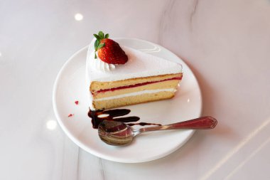 Beyaz kremalı bir dilim pasta ve beyaz bir tabakta duran bir çilek. Yanında bir çatal var. Pastada meyve dolgusu var gibi görünüyor, muhtemelen garnitürle eşleşen çilek..