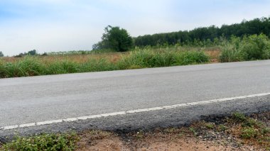 Mesafe boyunca uzanan boş asfalt yol. Beyaz çizgi yol kenarından geçiyor. Soldaki engebeli çimenli araziden ayırıyorum. Tarımsal alanların arka planı ve açık mavi gökyüzü.