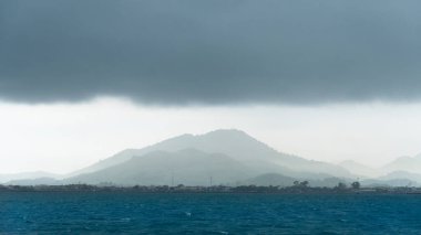 Siluetli dağ Khao Yai Da 'ya karşı kara yağmurlu gökyüzü ve önünde bir su kütlesi. Dağ puslu görünüyor. Mesafeyi gösteriyor. Kasaba ya da köyde belli belirsiz yapılar görülüyor..