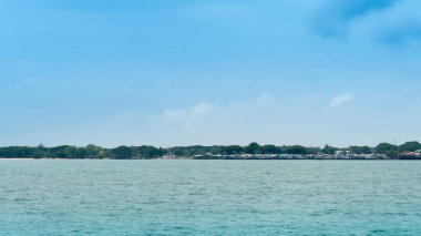 Ön planda deniz kıyısındaki şehirler. Sıraya dizilmiş ağaçlar var arkada. Deniz, resmin ön planındadır. Mavi gökyüzünün altında.