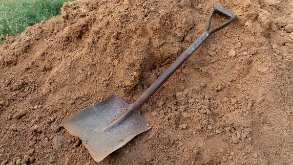 Çamur yığınının üzerinde yatan eski bir demir kürek. Yeşil bir çimenliğin yanında kahverengi bir demet toprak..